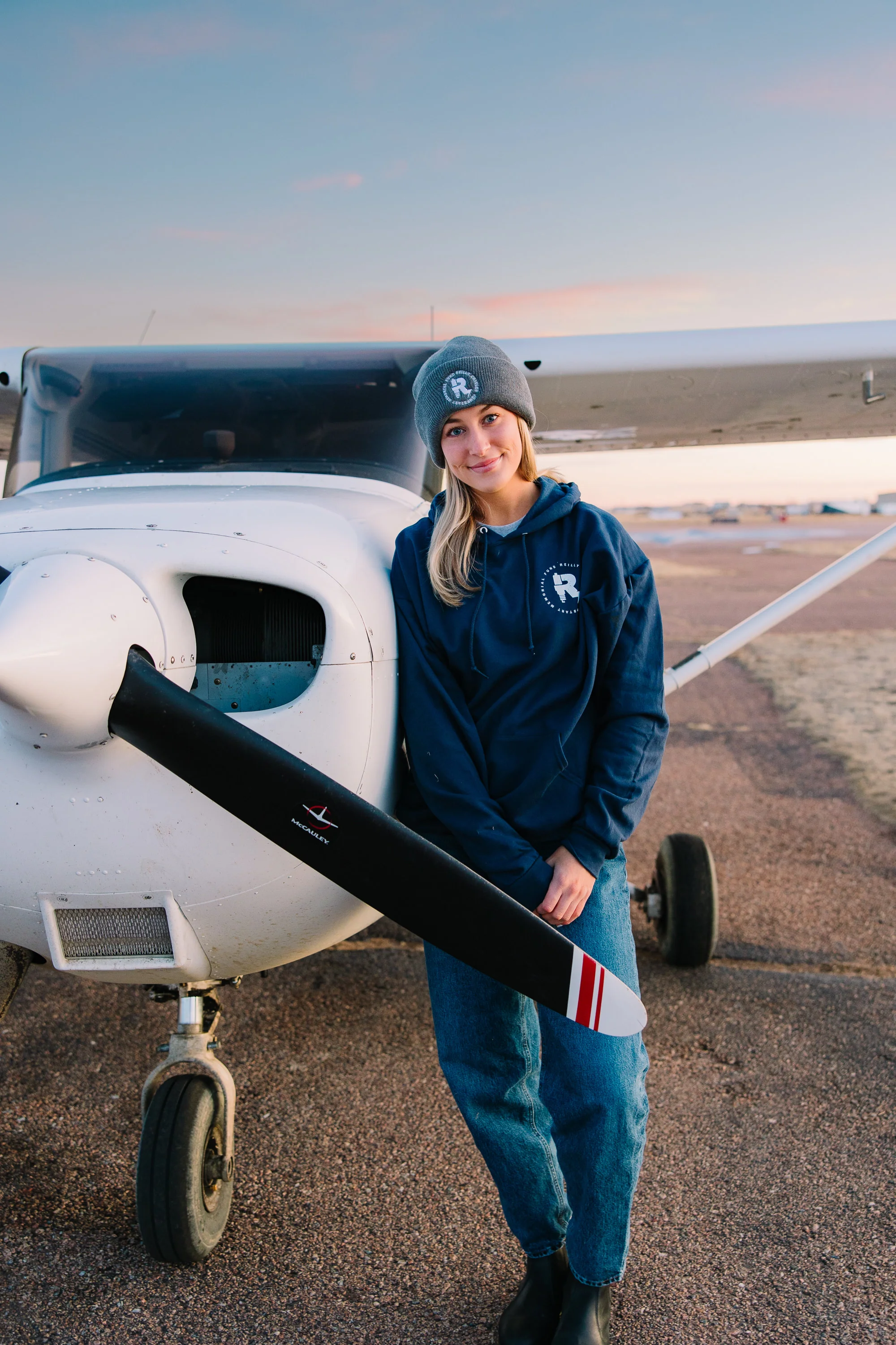 A young supporter in a Jumpstart R-monogram beanie and hoodie leans against a Cessna on the tarmac at dusk.