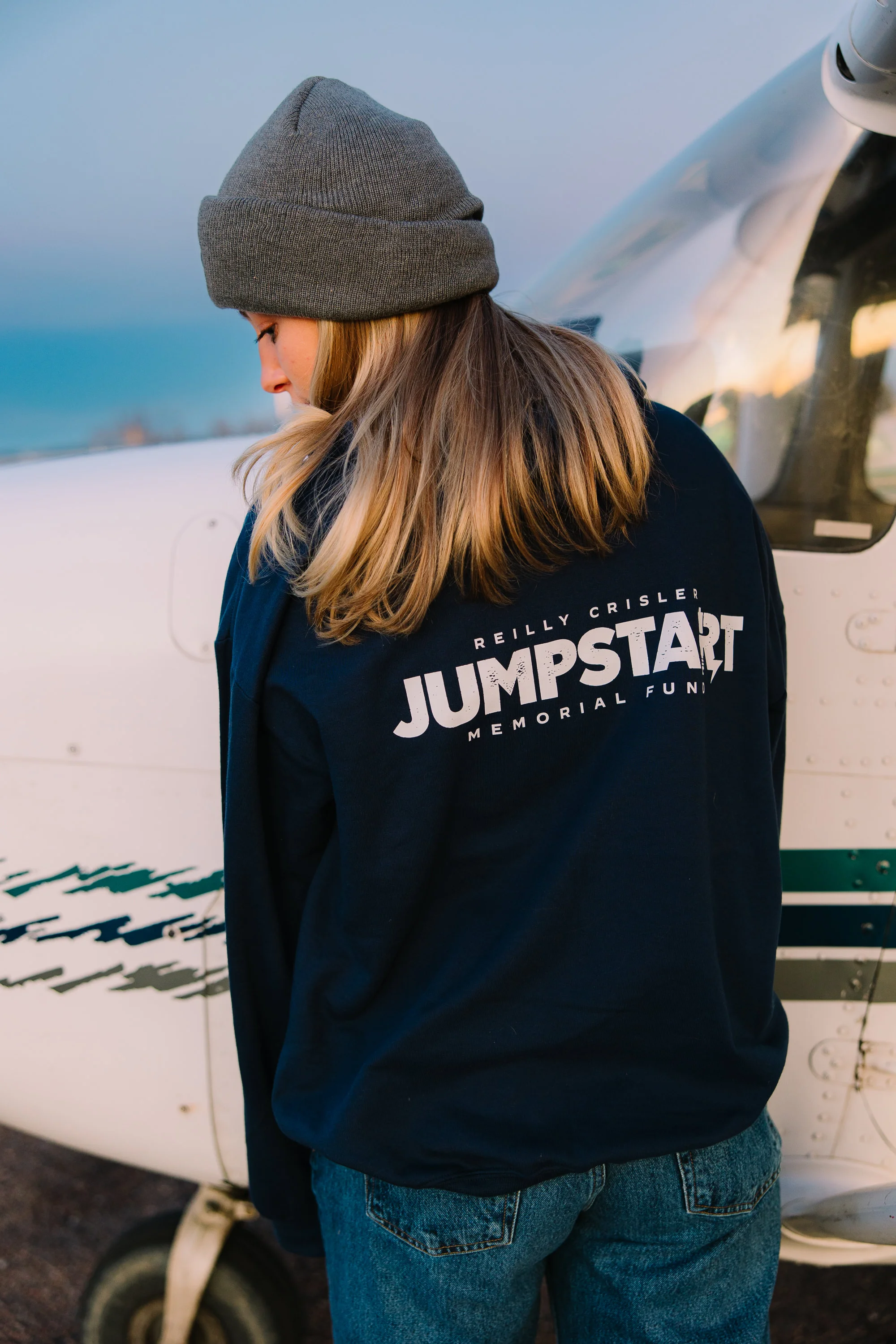 A young supporter wears a navy Reilly Crisler Jumpstart Memorial Fund long-sleeve beside a small aircraft at dusk.